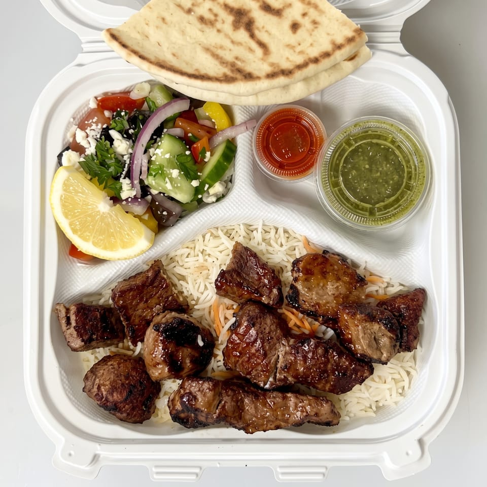 Grilled beef pieces over basmati rice with Greek salad, flatbread, lemon wedge, and red and green sauces.